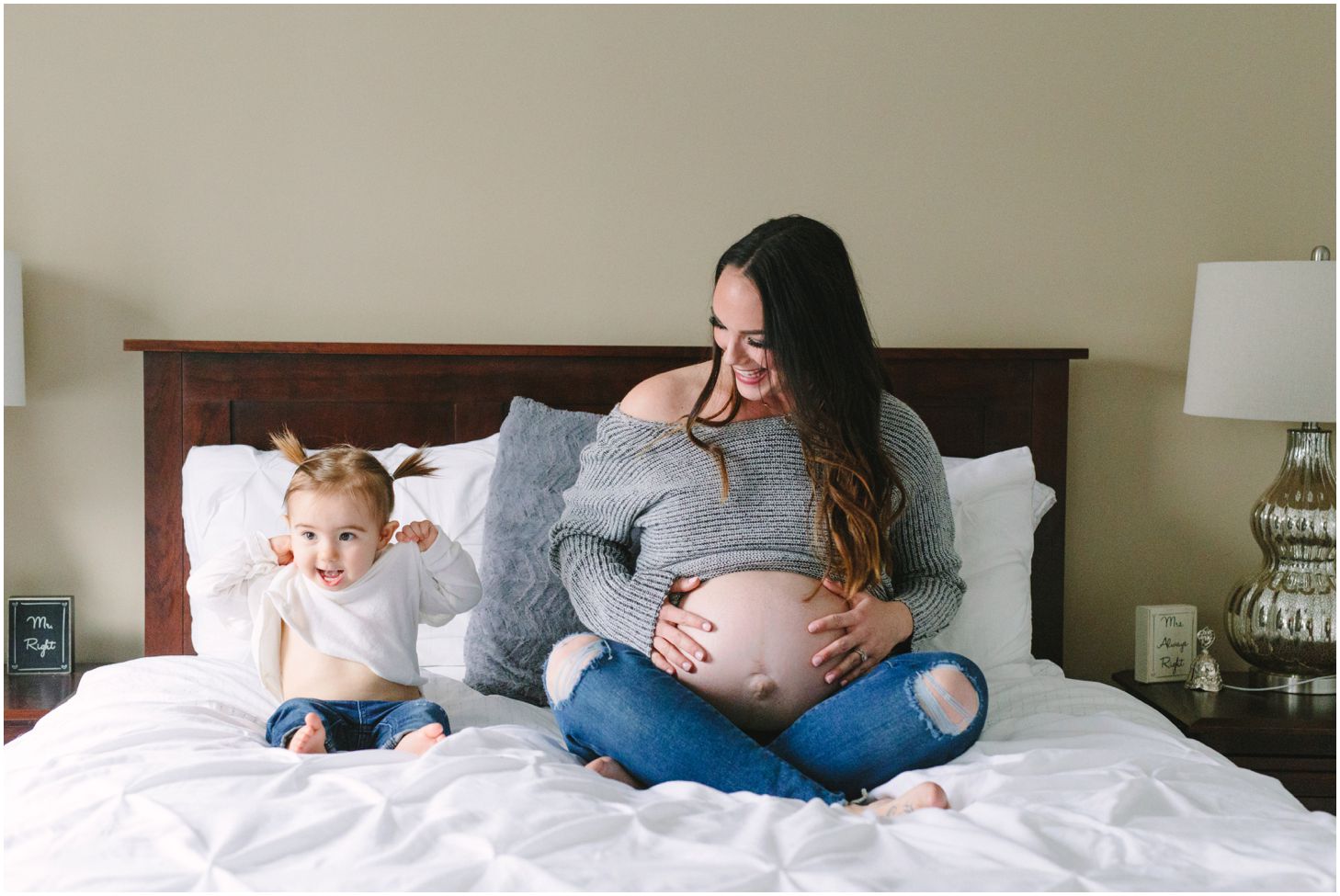 mom and little girl pose on a white bed together showing their bellies