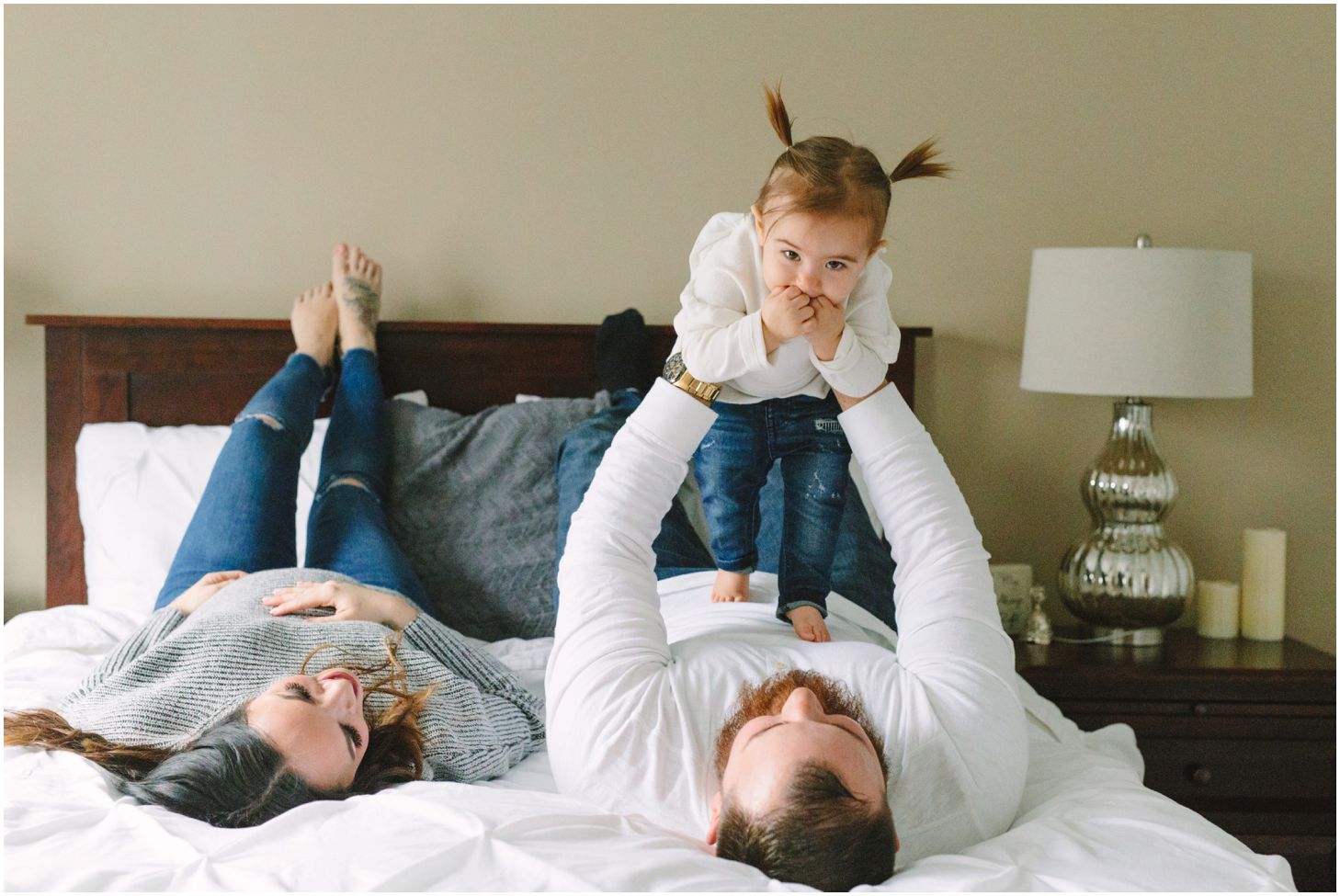 mom, dad, and little girl lay on a white bed together as dad lifts up the little girl