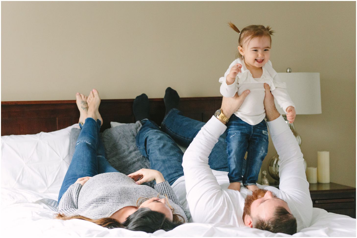 mom, dad, and little girl lay on a white bed together as dad lifts up the little girl
