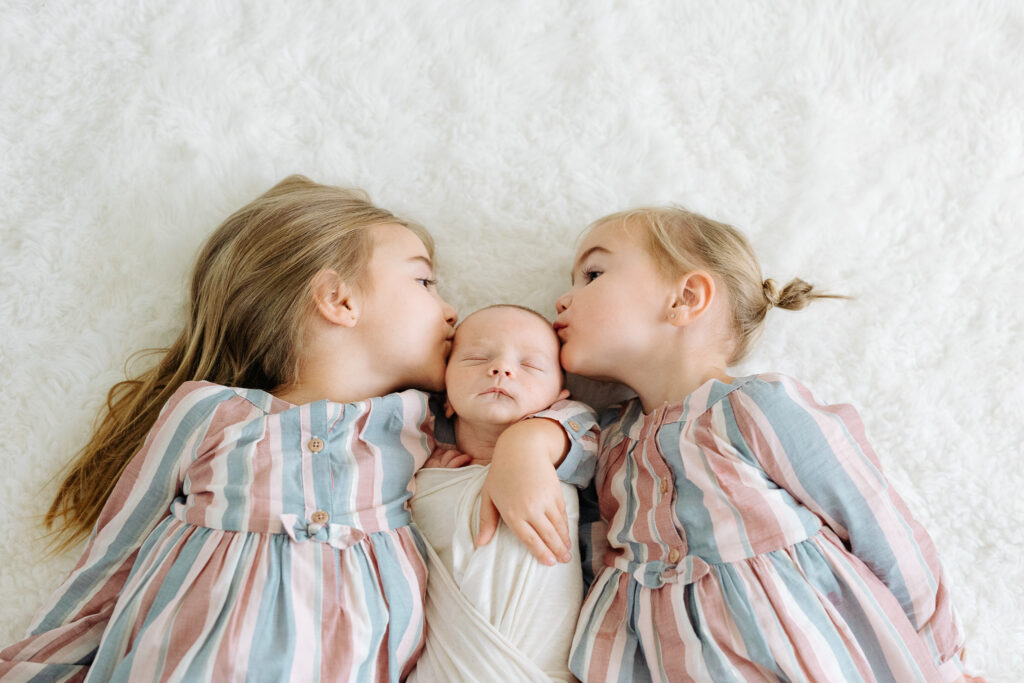 two girls in matching dresses kissing their newborn baby brother showing how to include siblings in a newborn photoshoot