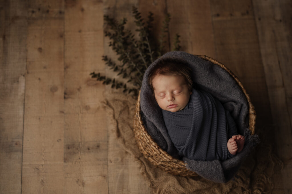 Adorable dark-haired baby boy in a slate blue wrap in a basket, captured by Kelly Adrienne Photography