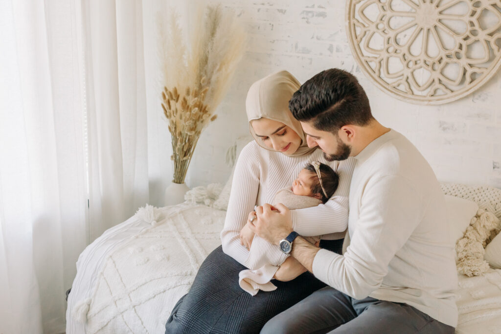 A sweet new family of three snuggling together on a neutral bed during their newborn session with Kelly Adrienne Photography