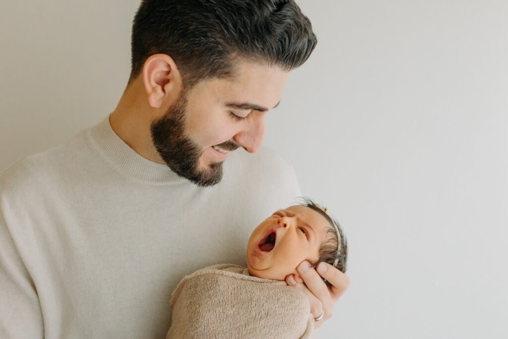 New dad smiling as he holds his yawning newborn baby girl, captured by Kelly Adrienne Photography
