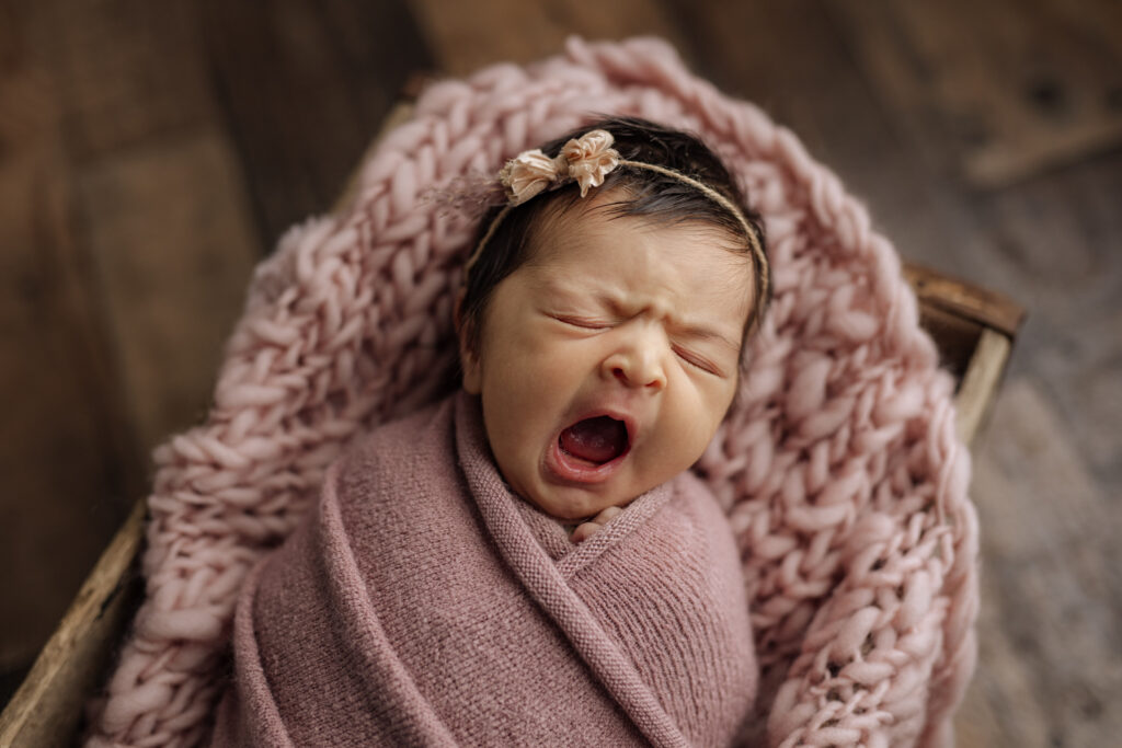 A yawning dark haired newborn girl in dusty pink at Kelly Adrienne Photography studio