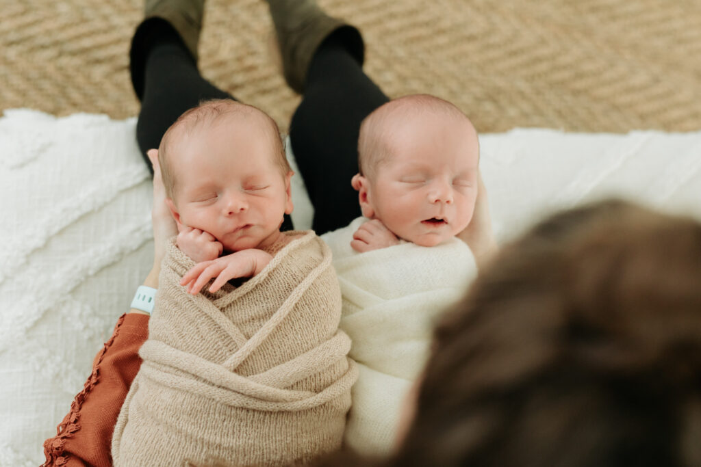 twin newborn boys in neutral wraps sleeping on mom's lap 
