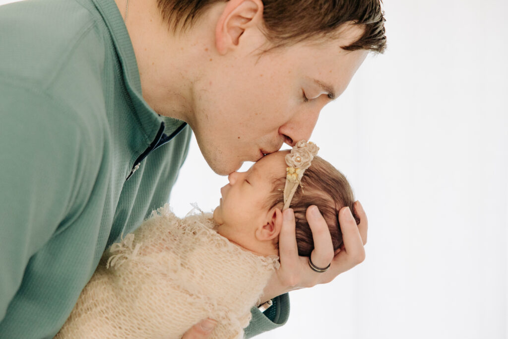 dad kissing his newborn baby girl in front of a window