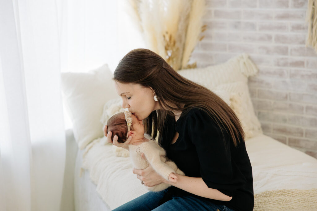 mom kisses her newborn baby while sitting on a bed in a boho studio