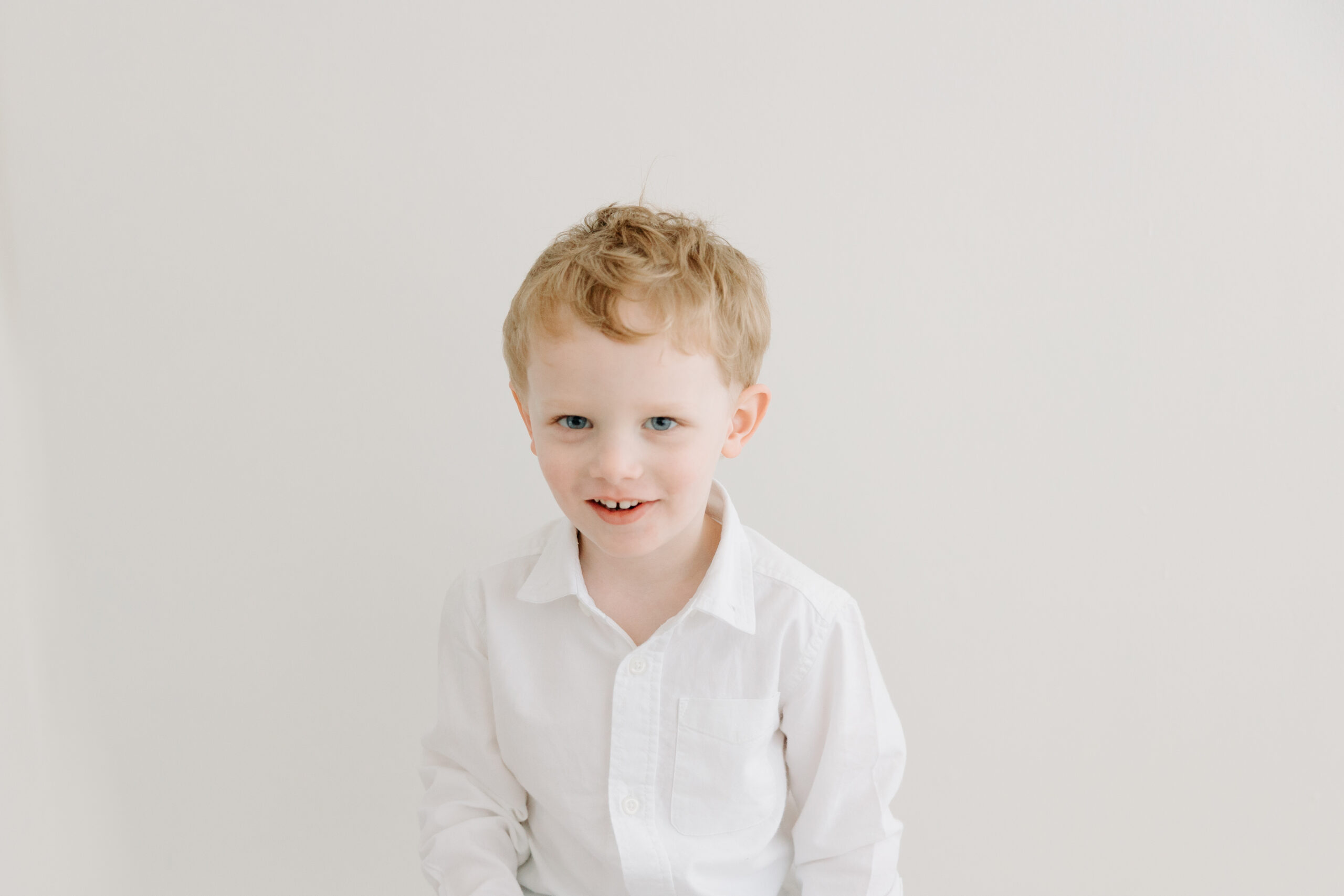 Newborn session with toddler | sweet toddler boy with blond hair smiles in front of a white wall