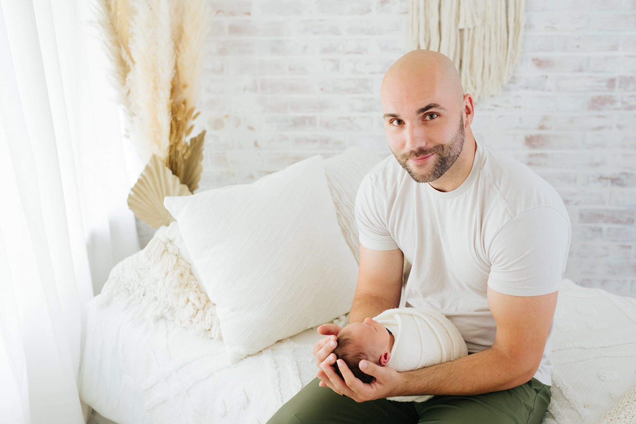 father holding his newborn baby while sitting on a bed in a white studio | Kelly Adrienne Photography