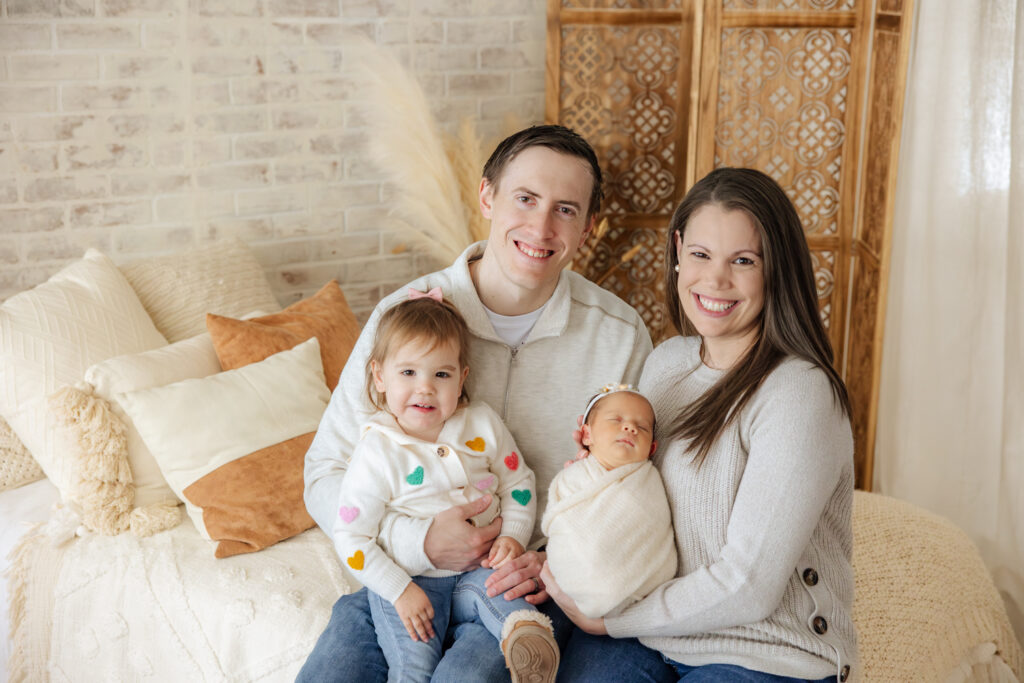 mom, dad, sister and newborn baby sitting on a white bed with a brick wall behind them, smiling