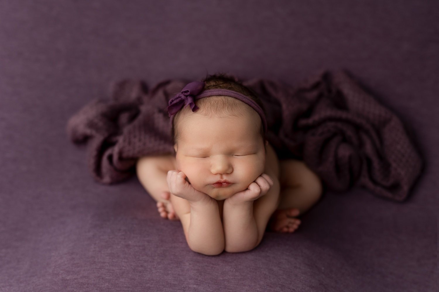 Newborn baby girl in the froggy pose with chin resting on hands against a purple backdrop