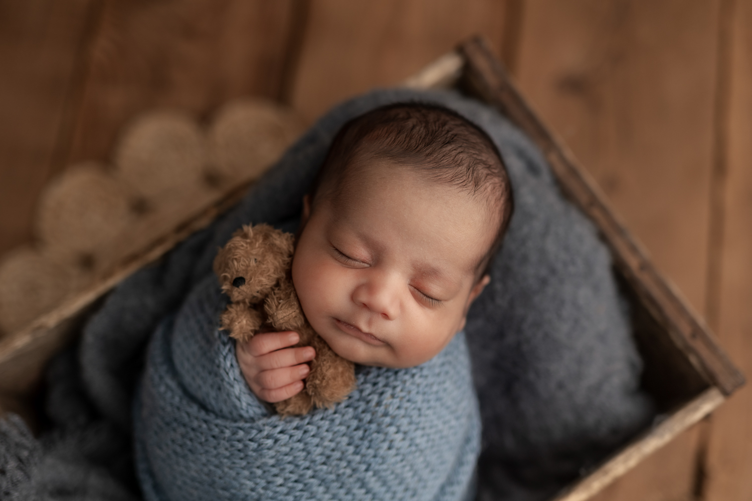 Newborn baby boy wrapped in a blue blanket, sleeping in a wooden crate and holding a teddy bear