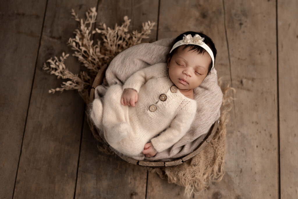 Sleeping newborn baby girl in cream pajamas with matching headband resting in a bowl with a wooden floor backdrop