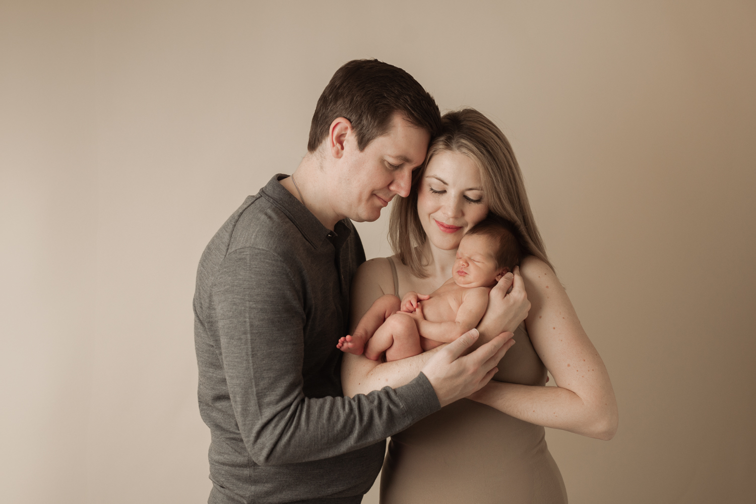 mom and dad snuggling with a newborn baby, standing in front of a neutral backdrop